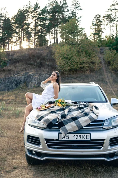 Attractive young woman is lying on the cars hood, enjoy a summer weekend picnic with a car outside the city in a field on a sunny day, sunset, vacation and trip.