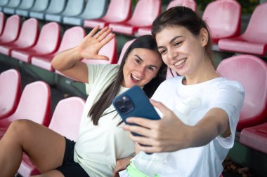 Stadyumdaki eğitimden sonra tribünde akıllı telefonla selfie çeken neşeli atletik genç kadınlar..