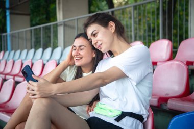 Stadyumdaki eğitimden sonra tribünde akıllı telefonla selfie çeken neşeli atletik genç kadınlar..