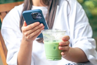 Matcha latte kahve içen ve dışarıdaki bir kafede akıllı telefon kullanan güzel genç bir kadın..