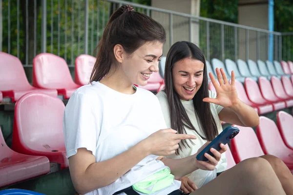 Fitness women sitting in the stands of the stadium and relaxing after workout, using smartphone.