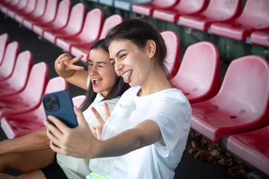 Stadyumdaki eğitimden sonra tribünde akıllı telefonla selfie çeken neşeli atletik genç kadınlar..