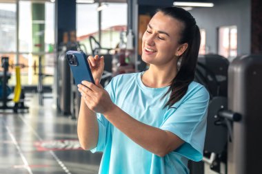 Spor salonunda cep telefonu kullanan genç bir sporcu, spor kıyafetlerini giyen bir kadın antrenmandan sonra telefona bakıyor, fitness merkezinde mesajlaşıyor..