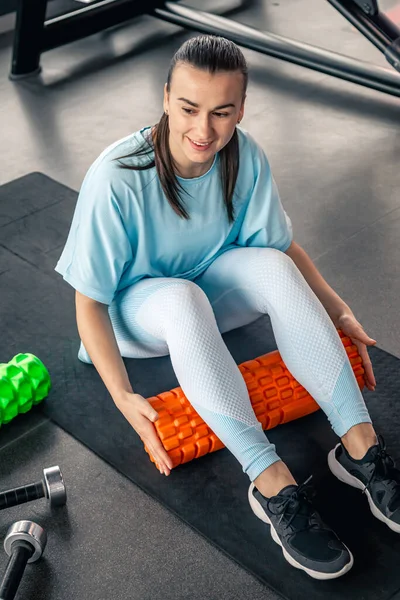 Happy woman works the fascia of the calf muscles with a massage foam roller on the floor in a gym.