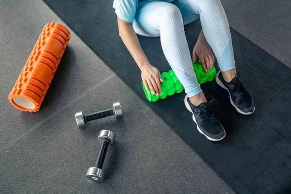 Woman works the fascia of the calf muscles with a massage foam roller on the floor in a gym.