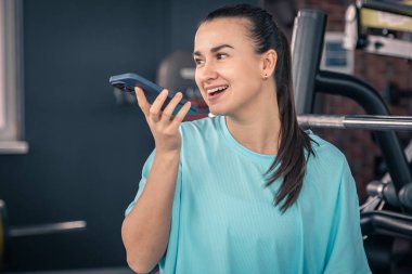 Sporcu kadın spor salonunda elinde akıllı telefonuyla biriyle konuşuyor ya da sesli mesaj kaydediyor..