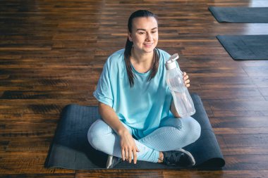 Mavi spor giysili genç bir kadın antrenmandan, yogadan ve sağlık için esneme hareketlerinden sonra mindere oturup su içiyor..