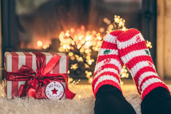 Feet in Christmas socks and gift box in front of the fireplace, close-up, relaxing at home, cozy background, copy space.