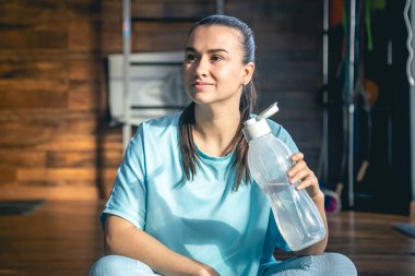 Mavi spor giysili genç bir kadın antrenmandan, yogadan ve sağlık için esneme hareketlerinden sonra mindere oturup su içiyor..
