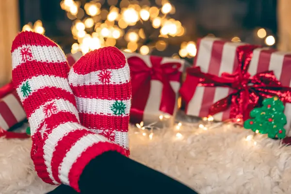 Feet in Christmas wool socks near fireplace with gift boxes in winter time, resting at home, copy space.