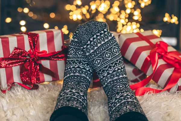 Feet in Christmas socks and gift boxes in front of the fireplace, close up, relaxing at home, cozy background.