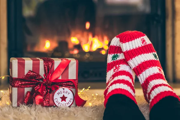 Feet in Christmas socks and gift box in front of the fireplace, close-up, relaxing at home, cozy background, copy space.