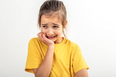 Sarı tişörtlü, korkmuş ve endişeli küçük bir kızın portresi izole edilmiş beyaz bir arka planda, tırnaklarını yiyor, insan duyguları kavramı..