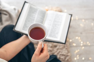 Kadın ellerinde bir fincan çay, yakın plan. Bir kadın yerde, halının üzerinde otururken kitap okuyor, boşluğu kopyalıyor..