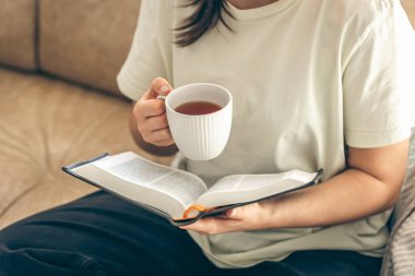 Genç bir kadın evde kitap okuyor, kanepede dinleniyor. Yakın plan, kitap ve fincan çay kadınların elinde..