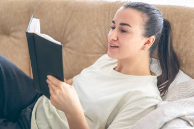 Genç bir kadın evinde, elinde bir kitapla kanepede uzanıyor. Sabah yaşam tarzı konsepti.