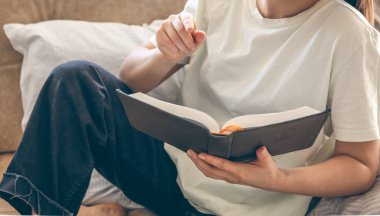 Evde kitap okuyan genç beyaz bir kadın. Bir kadın kanepede kitap okuyor. Hobiler, ev eğlencesi ve iyi bir okuma alışkanlığı..