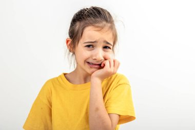 Sarı tişörtlü, korkmuş ve endişeli küçük bir kızın portresi izole edilmiş beyaz bir arka planda, tırnaklarını yiyor, insan duyguları kavramı..