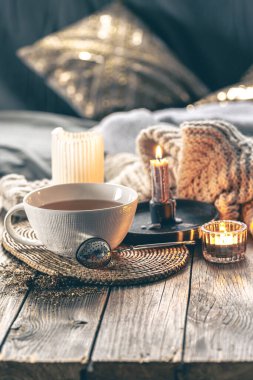 Sıcak ev kompozisyonu, bir fincan çay, yanan mumlar ve odanın arka planında örülmüş bir element..