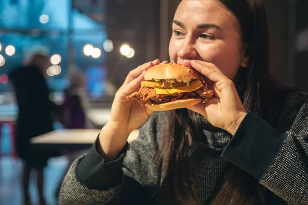 Attractive young woman eating a chicken burger in a fast food cafe. Unhealthy high-calorie food and fast food, copy space.
