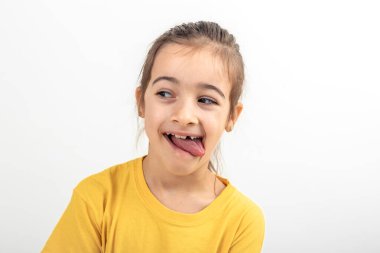 Sarı tişörtlü küçük bir kız, dili beyaz bir arka planda sallanırken yüzünü buruşturuyor..