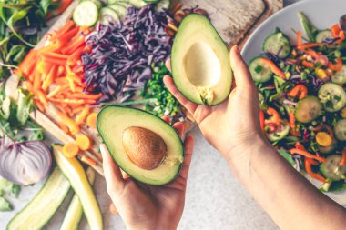 Kesik bir avokadonun kadın ellerinde yakın plan çekimi. Bir kadın iki parçaya ayrılmış bir avokado tutar. Taze sebzelerden salata hazırlıyorum. Üst görünüm.