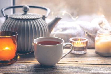 Bir fincan çay, mumlar ve çaydanlıkla rahat bir ev kompozisyonu. Pencere kenarındaki ev rahatlığı. Boşluğu kopyala.