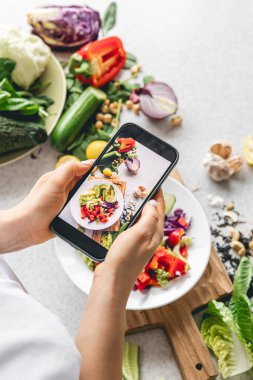 Bir kadın mutfak masasında taze sebze salatası fotoğrafı çekiyor. Bir blogcu içerik yaratır. Bir yemek fotoğrafçısı blog için fotoğraf çekiyor..