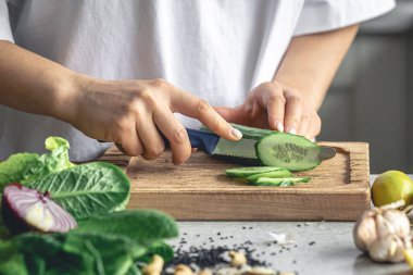 Bir kadın mutfaktaki tahta bir tahtanın üzerinde taze salatalık kesiyor. Vejetaryen sebze salatası hazırlıyorum. Yakın plan, sebze doğrayan eller..