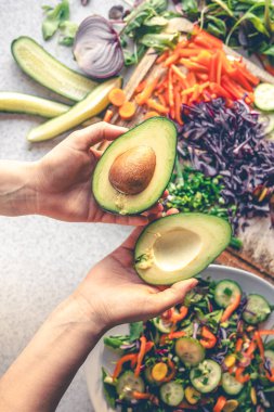 Kesik bir avokadonun kadın ellerinde yakın plan çekimi. Bir kadın iki parçaya ayrılmış bir avokado tutar. Taze sebzelerden salata hazırlıyorum. Üst görünüm.