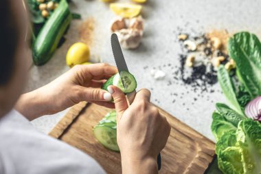 Bir kadın mutfaktaki tahta bir tahtanın üzerinde taze salatalık kesiyor. Vejetaryen sebze salatası hazırlıyorum. Yakın plan, sebze doğrayan eller..