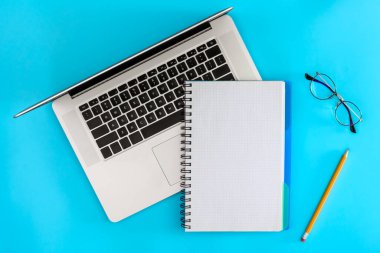 Boş bir defter, dizüstü bilgisayar ve mavi arka planda gözlük, üst manzara. Planlama ve eğitim konsepti. Minimalist mavi ofis arkaplanı.