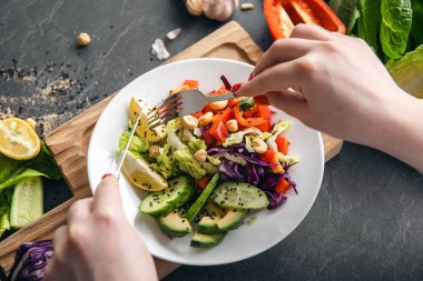 Bir kadın taze sebzelerden vejetaryen salatası yiyor, siyah bir mutfak masasında yakın plan bir tabak. Sağlıklı beslenme konsepti. Evde yemek pişirmek..