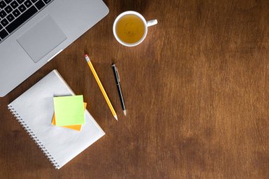 Laptop, bir fincan çay ve ahşap bir masada boş not defteri, üst manzara. Düz ofis masası, kopyalama alanı.