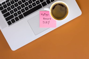 Laptop klavyesine Mutlu Patronlar Günü ve kahverengi ofis masasına bir fincan kahve iliştirilmiş yapışkan bir not..