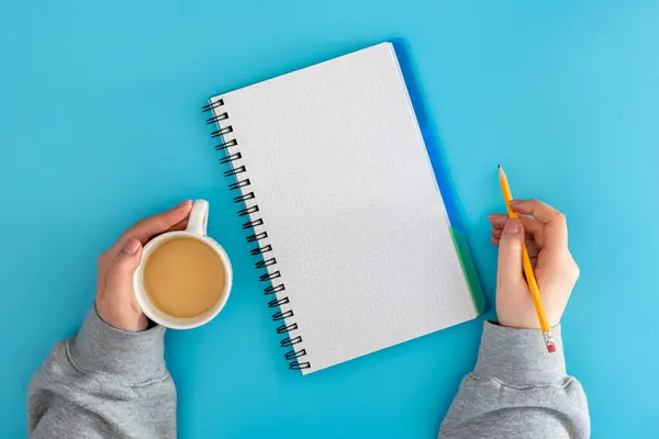 Woman with a cup of coffee writes in a notepad on a blue background, top view. Planning concept. Ideas for business.
