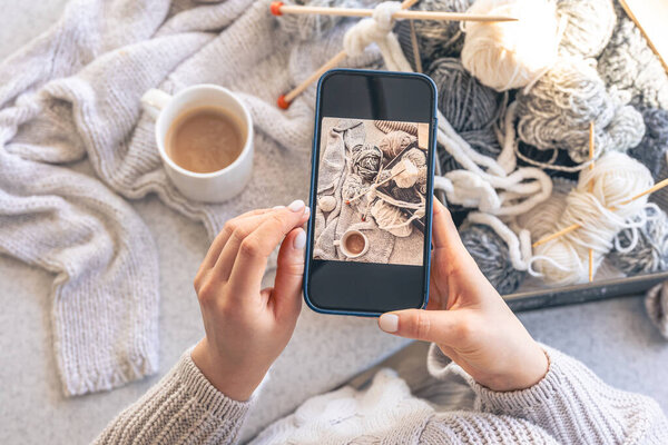 A woman takes pictures of yarn, threads and a cup of coffee on her smartphone. Home hobby concept.