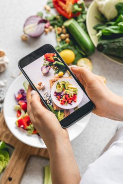 Bir kadın mutfak masasında taze sebze salatası fotoğrafı çekiyor. Bir blogcu içerik yaratır. Bir yemek fotoğrafçısı blog için fotoğraf çekiyor..