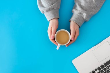 Kadın ellerinde bir fincan kahve ve mavi arka planda bir dizüstü bilgisayar. Kopyalama alanı olan üst görünüm, düz yatay.