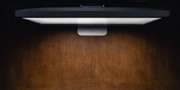Computer monitor on a wooden table in a dark room, top view. Night work concept. Copy space.