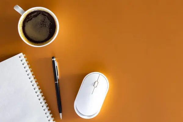 Notepad, pen, computer mouse and cup of coffee on a brown background, flat lay, space for text.