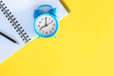 Sarı zemin üzerinde mavi alarm saati ve not defteri, minimal arkaplan, düz tasarım, kopyalama alanı.