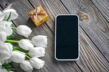 Akıllı telefonlu bahar arka planı, sarı hediye kutusu ve ahşap arka planda bir buket beyaz lale çiçeği, üst manzara..