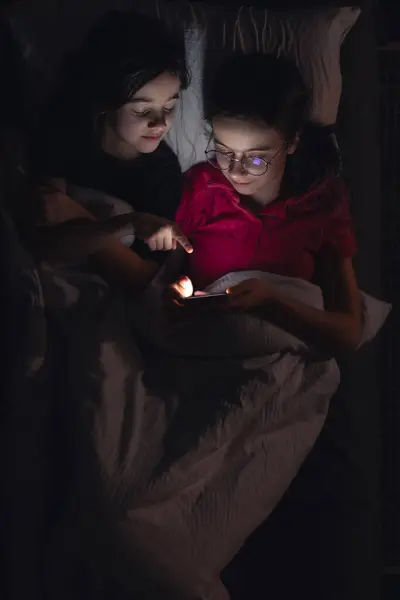 Teenage girls with a smartphone lies in bed at night, top view. Children using smartphone lying in bed late at night, playing games, watching videos online, scrolling screen.