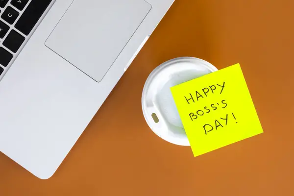 Sticky note with phrase Happy Boss Day attached to cup of coffee on brown office table, top view.