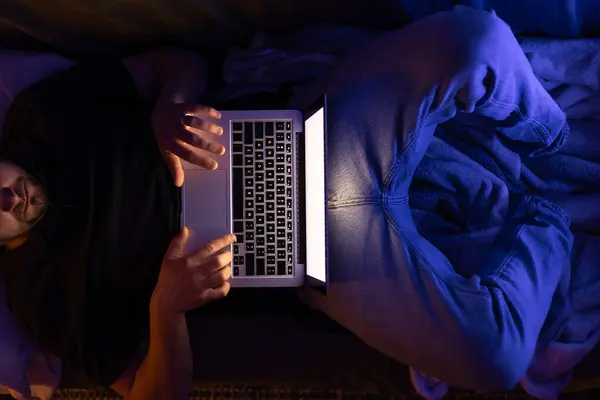 Young businessman or freelancer working overtime with laptop at home, lying on sofa late at night, has deadline at work. Insomnia and computer addiction.