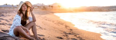 Güneşin batışında, kafasında şapka olan beyaz bir mayo giymiş güzel bir manken kumsalda poz veriyor. Web, sosyal medya afişi şablonu. Boşluğu kopyala.