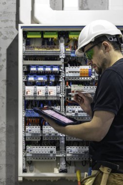 Elektrik santralinde fitillerle çalışan bir elektrik teknisyeni. Elektrik ekipmanlarının kurulumu ve bağlantısı. Profesyonel bir tablet kullanır.