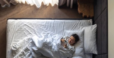 Genç bir kadın yatağa uzanıyor ve akıllı telefon kullanıyor. Sabahları yatakta telefonlu bir kadın. Otelde dinlenen bir kadın, tembel bir sabah. Yukarıdan görüntüle.