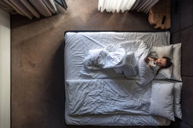 Genç bir kadın yatağa uzanıyor ve akıllı telefon kullanıyor. Uyan ve rahat bir yatak odasında cep telefonunda internete göz at. Otelde dinlenen bir kadın, tembel bir sabah. Yukarıdan görüntüle.